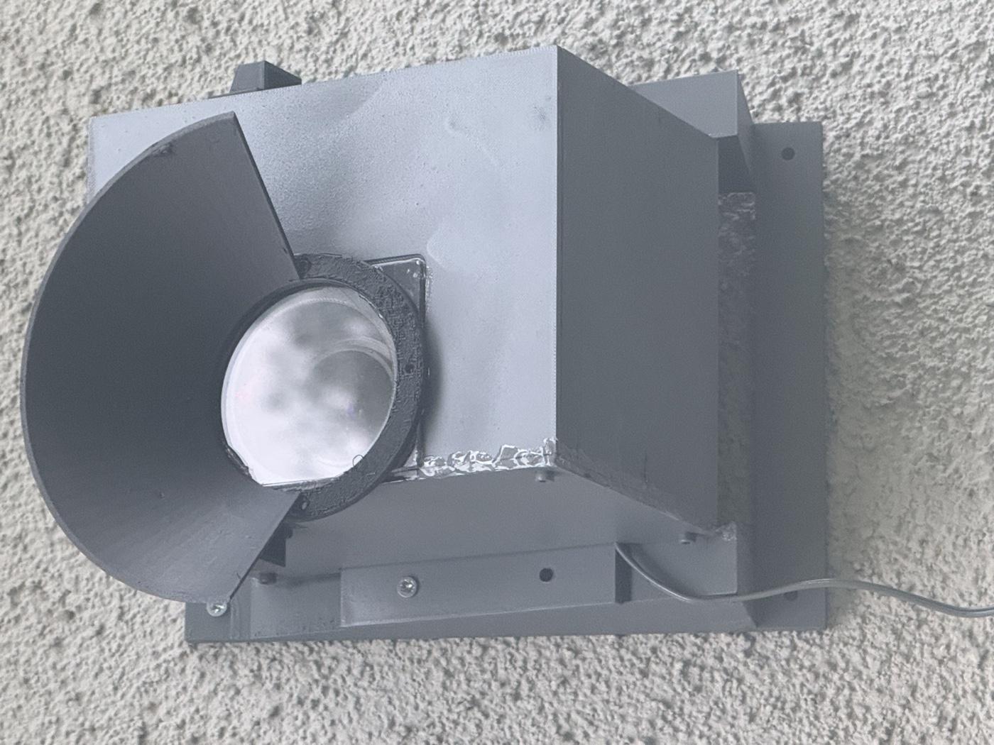 The finished gray enclosure mounted on the stucco wall, sun visor over the windowed lens
